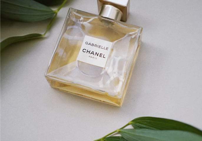 A glass bottle of Gabrielle Chanel perfume sits on a white surface surrounded by green leaves